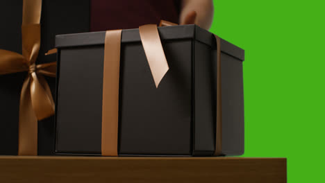 Close-Up-Of-Man-Picking-Up-Gift-Wrapped-Present-In-Box-Decorated-With-Ribbon-On-Table-Shot-Against-Green-Screen